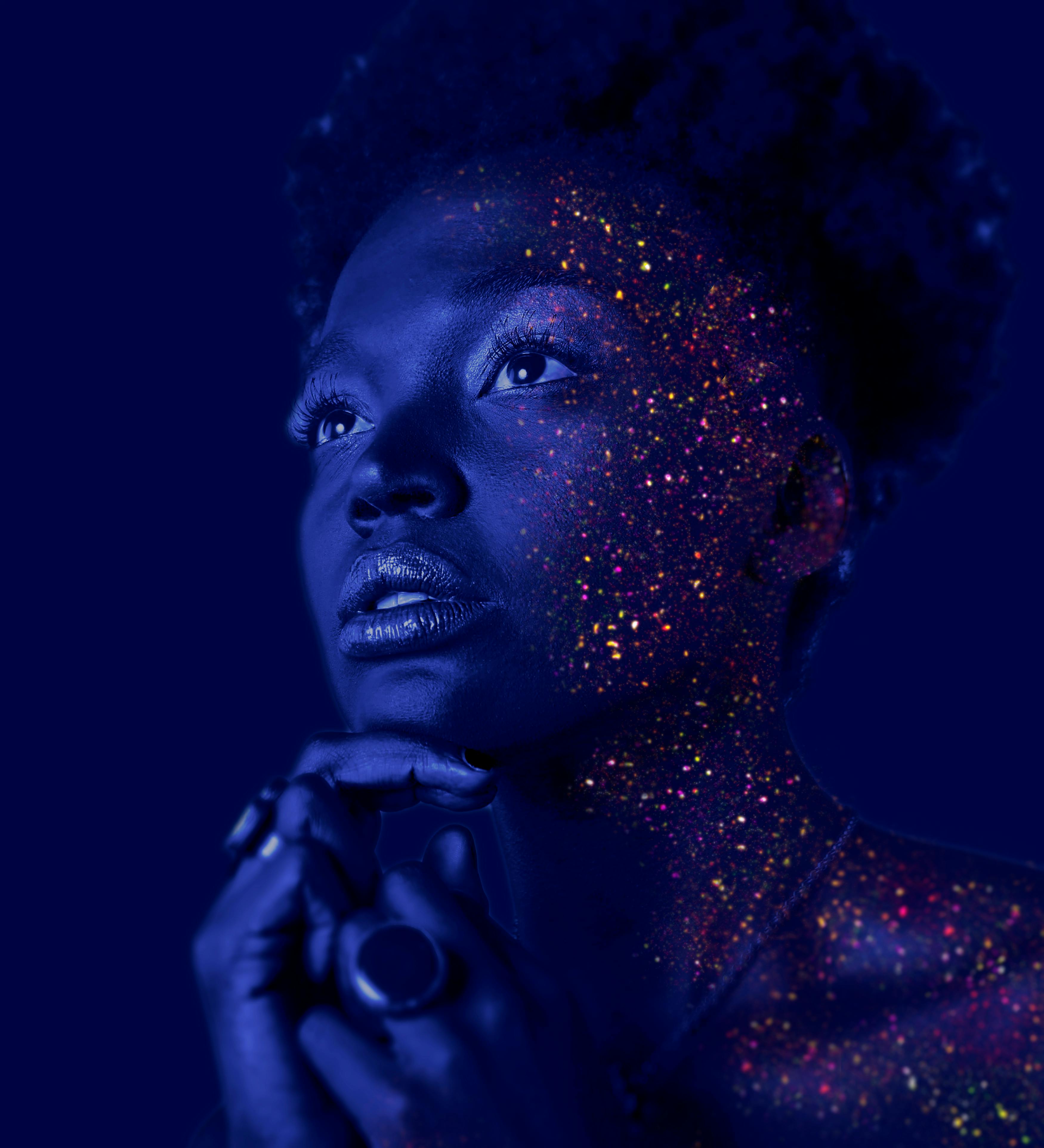 image of a black woman staring off with a blue futuristic filter and multicolored glitter painted on her face and shoulder