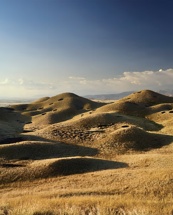 Rolling sand hills in a dessert with mountains in the back. The sun is shining down and reflecting off the hills. 