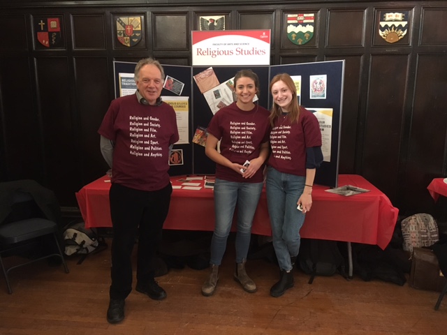 Three people standing in front of a School of Religion poster