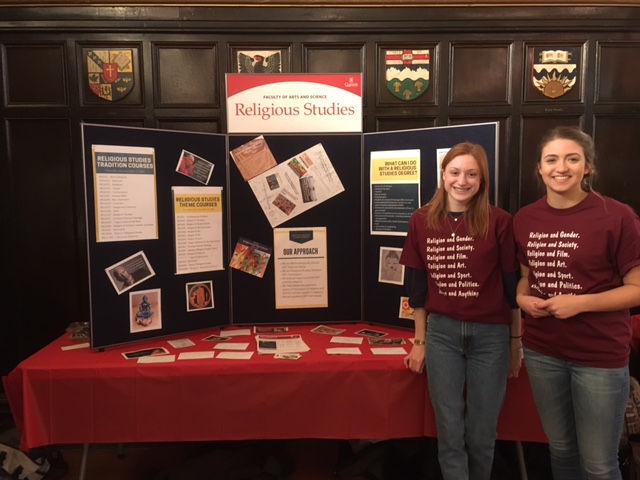 Two people smiling in front of a School of Religion poster