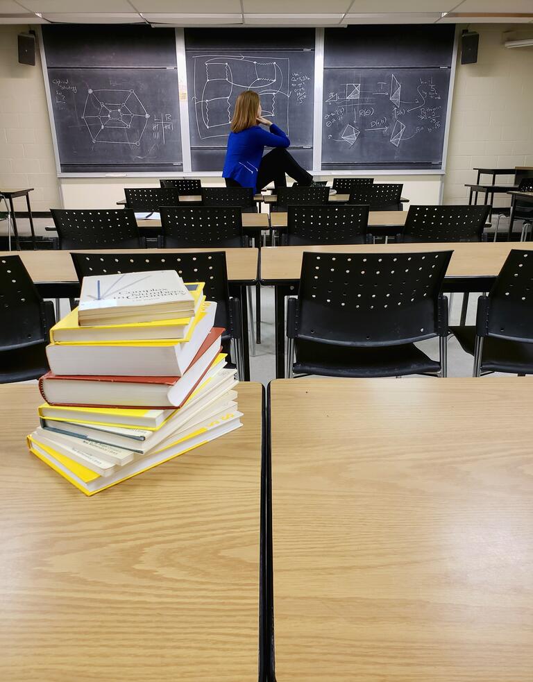 Woman sitting in front of chalk board with books stacked on a desk