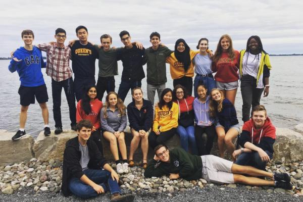 Group of students standing together smiling on the rocks in front of the Kingston waterfront. 