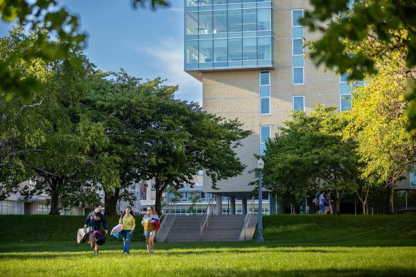 Three students walk forward, surrounded by greenery, and a residence building in the background.
