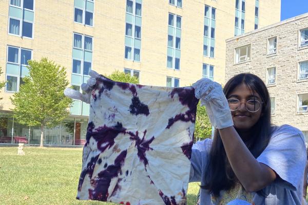 A student is wearing gloves and is holding up a tie-dyed fabric on a sunny day outdoors.