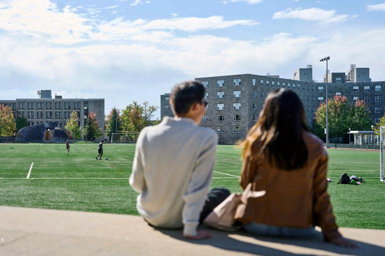 Two students sit with their backs to the camera and face Tindall Field on a sunny day.