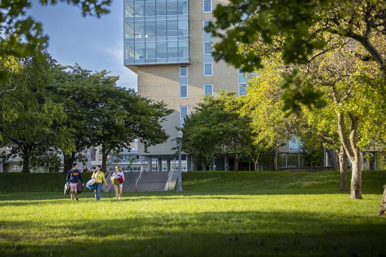 Three students walk on a grassy area with trees. Brant House is in the background.