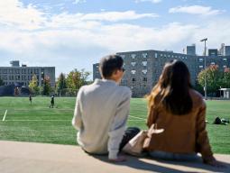 Two students sit with their backs to the camera and face Tindall Field on a sunny day.