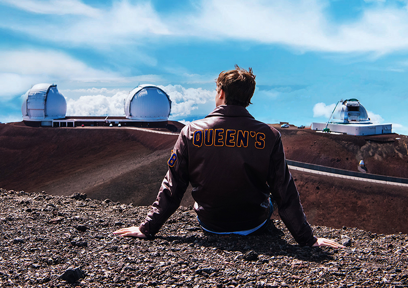 A student wearing a Queen's jacket sits on a mountaintop overlooking several observatories.