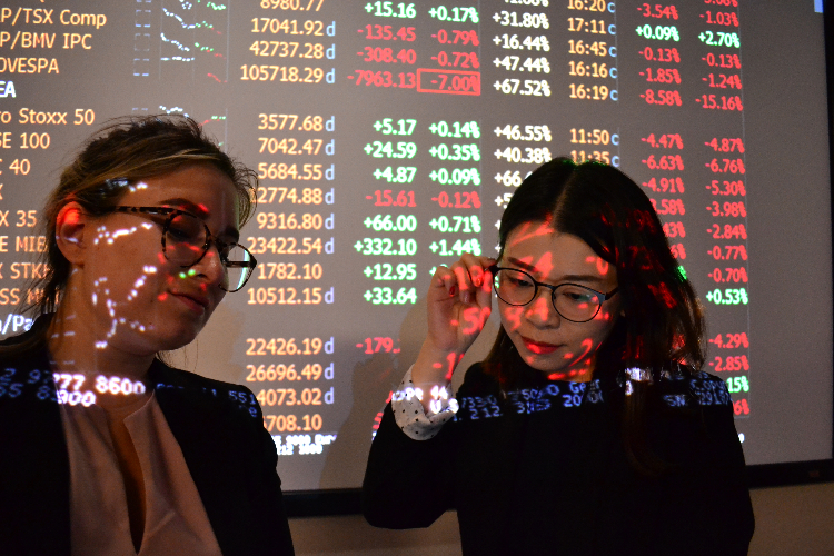 [Two students stand in front of a stock ticker board].