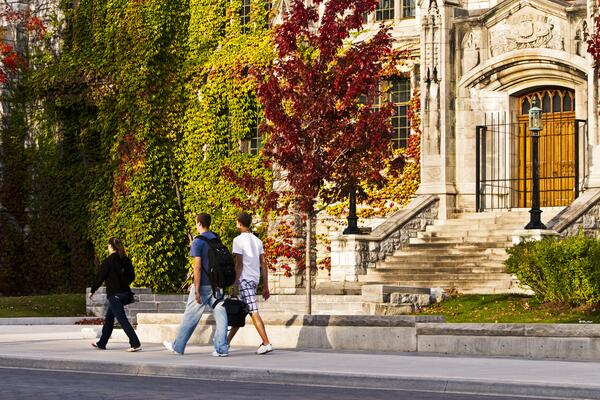 three students walking across campus in front of a limestone building and fall coloured trees