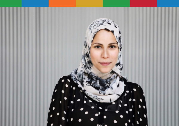 Woman with white and grey watercolour hijab and a black shirt with white polka dots standing in front of a grey wall