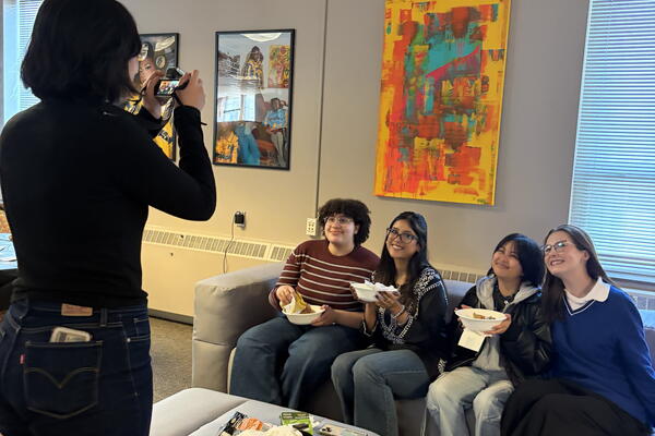Student standing while taking a picture of four students seating on a couch.