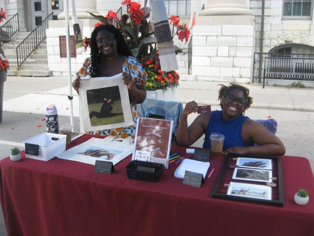 two black women at a red table displaying artistic tote bags